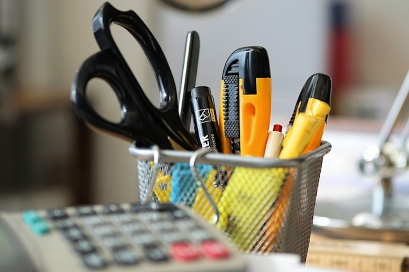 Taschenrechner neben Stifte-Becher mit Schere, Cutter und anderen Büroartikeln.