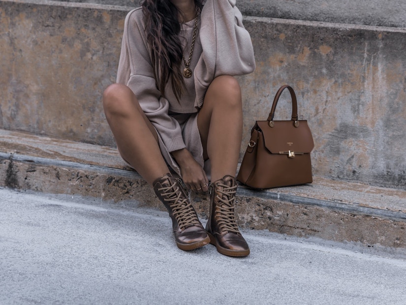 Auf einer Betonfläche sitzt eine Frau mit braunen Stiefeln neben einer braunen Lederhandtasche