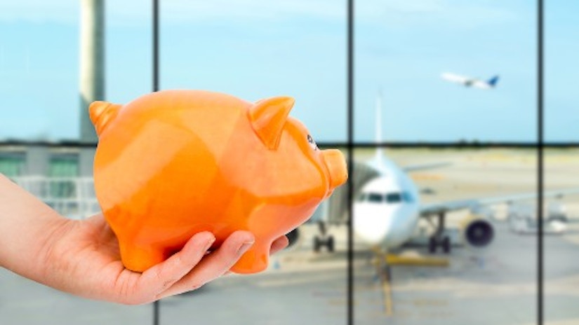 Eine Hand hält ein Sparschwein. Im Hintergrund sieht man Flugzeuge.