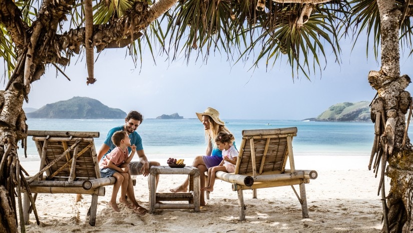 Eine Familie sitzt am Strand auf Holzpritschen unter Palmen und lacht.