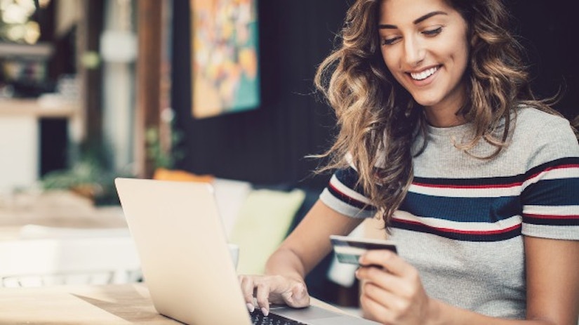 Eine Frau sitzt am Computer, guckt auf ihre Kreditkarte und lächelt.
