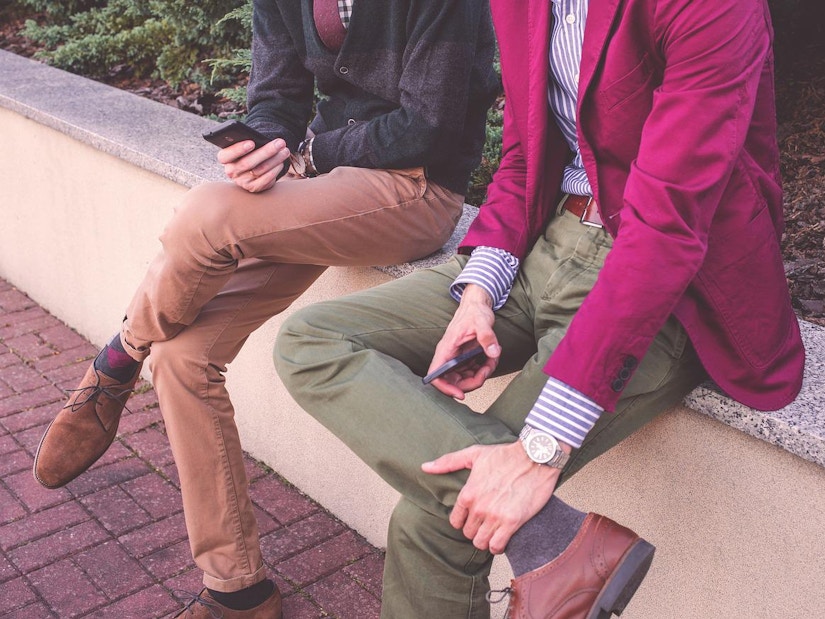 Zwei Männer: einer in brauner Hose und dunkelblauem Pullover, der andere in einer rosa Jacke