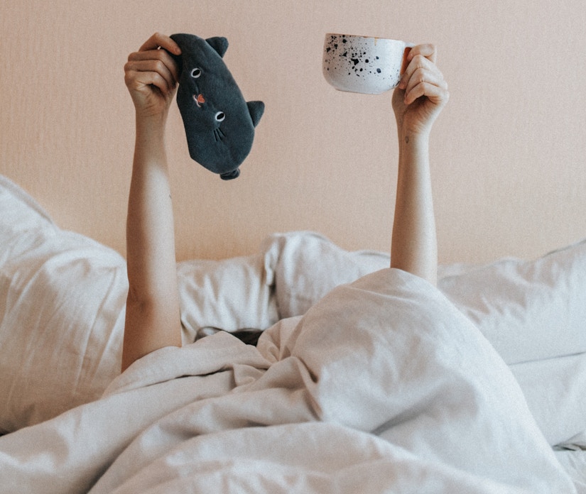 Person im Bett hält Kaffeetasse in die Höhe