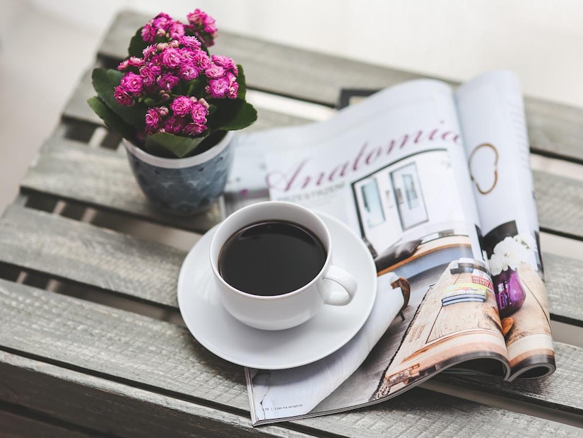 Eine kleine Vase mit Blumen, eine Tasse Kaffee und eine Zeitschrift liegen auf einem Holztisch 