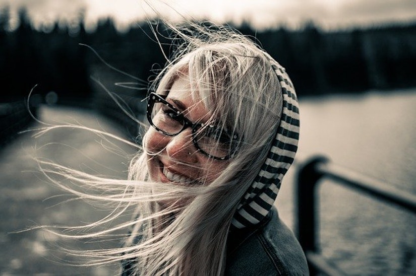 Blonde Frau bei Wind am Wasser mit Jacke, gestreiftem Kapuzenpulli und Brille mit dunklem Rand.
