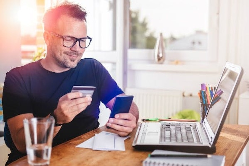 Ein Mann sitzt mit Kreditkarte und Smartphone vor seinem Laptop und lächelt