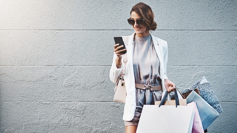 Eine Frau hat Einkaufstüten in der Hand und schaut lächelnd auf ihr Handy.