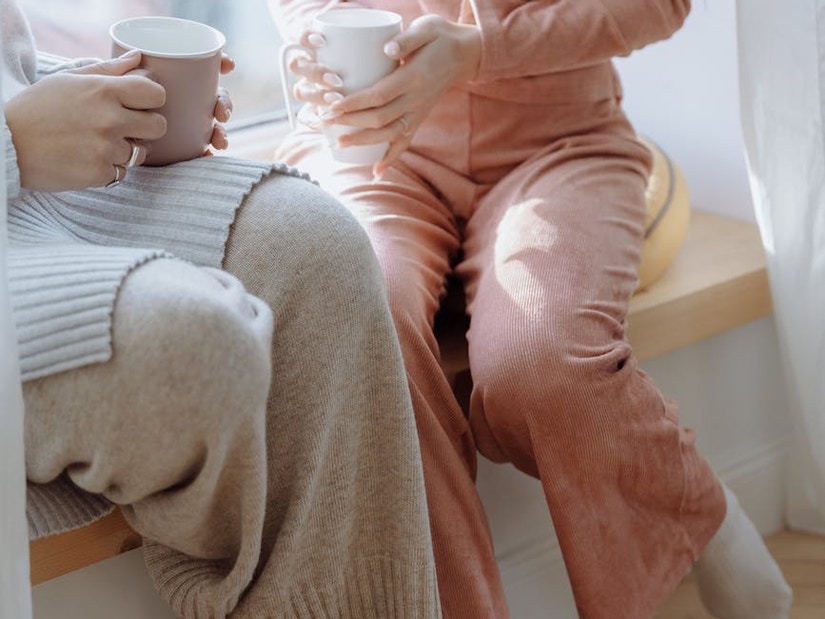 Zwei Frau sitzen in Pyjamas an der Fensterbank und trinken heißen Kaffee